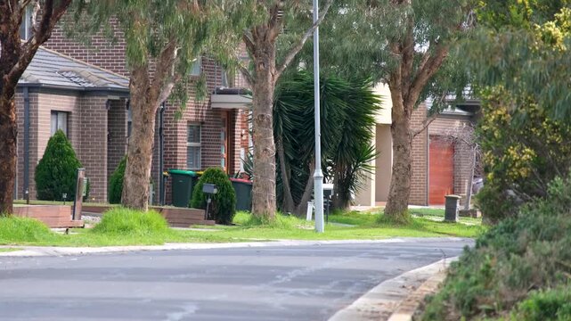 A typical new housing estate street with tall drought-tolerant native gum trees, an unkempt nature strip, and artificial turf front yards outside brick houses in an outer suburb of Melbourne Australia