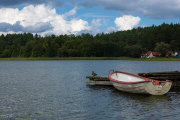 Weathered white and red rowboat moored beside a rustic wooden dock, with a mallard duck, forest and lakeside houses under dramatic clouds. © Do