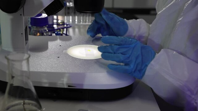 Scientist in protective suit using microscope in laboratory. Close-up of a scientist in a cleanroom suit carefully placing a specimen on a microscope slide using tweezers for scientific research.