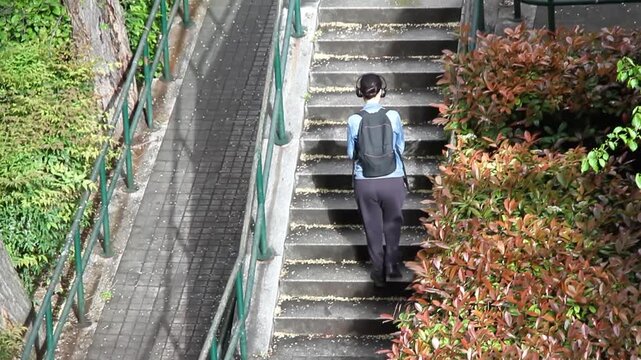 Mujer subiendo escalera, pelda&ntilde;o y escal&oacute;n paseo con cascos de m&uacute;sica, mochila y cazadora vaquera upstairs climb step woman por la calle con barandilla,  adoqu&iacute;n y arbusto de seto y matorral