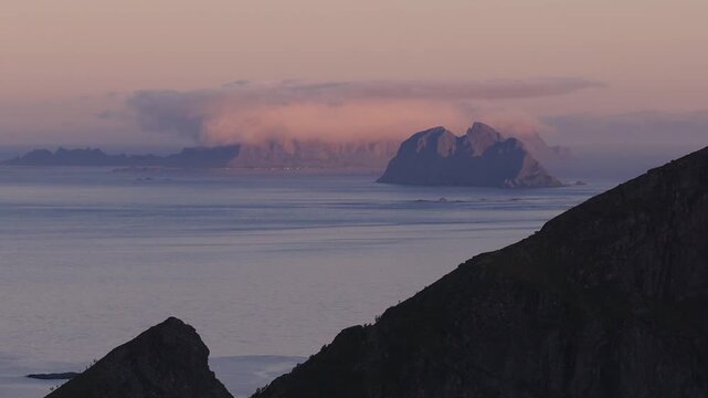 An evening view over the Norwegian Sea reveals distant mountain islands under pink clouds at dusk in Lofoten, Norway.