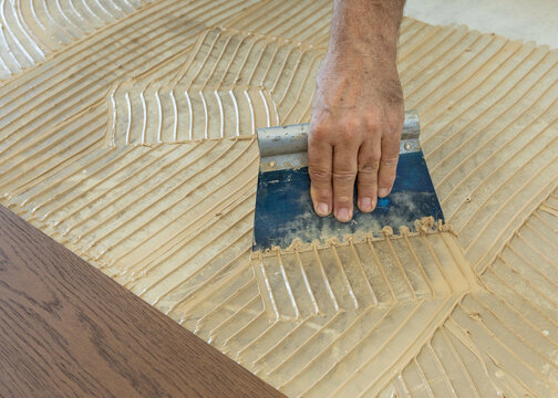 Worker applying adhesive on concrete floor with notched trowel during oakwood parquet installation