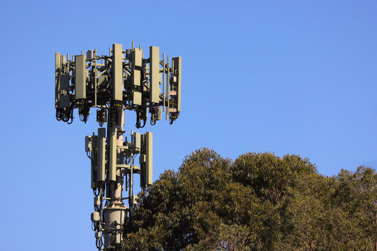 View of a 5G tower with telecommunications equipment and antennas set against a clear blue sky and trees in Sydney, New South Wales, Australia.