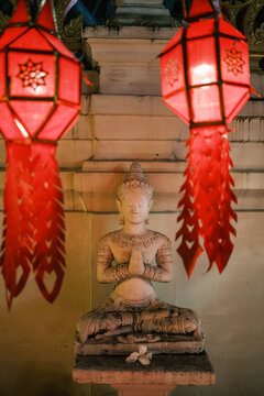 View of a serene stone Buddha statue in a prayer pose, framed by two glowing red traditional paper lanterns in a temple setting, Chiang Mai, Thailand.