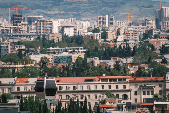 View of the cable car moving above the cityscape of Tbilisi with historic and modern buildings, lush trees, and construction cranes in the background in Tbilisi, Tbilisi, Georgia.