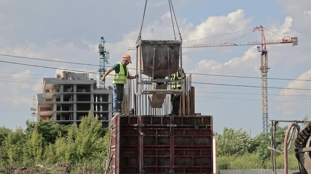 Construction workers guide suspended concrete hopper above reinforced formwork at building site, Tower cranes and unfinished structure frame active concrete work in daylight.