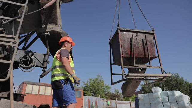 Construction worker guides concrete hopper suspended beside mixer truck at building site, Heavy machinery and lifting chains support active concrete handling outdoors.