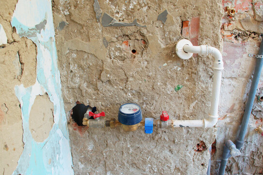 Water meter and exposed plumbing pipes installed on an old damaged wall during home renovation. Close-up of a water supply system with valves, pipe fittings and rough plaster. The unfinished wall.