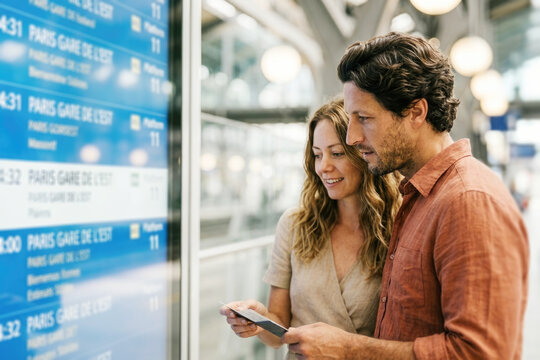 Happy tourist couple checking train schedule timetable board at railway station