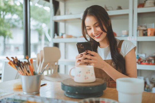 Smiling woman using smartphone while working with pottery in creative ceramic studio