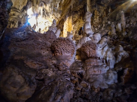 Massive stalactites and detailed rock formations fill this subterranean limestone cavern featuring textured stone pillars illuminated by natural light filtering through the deep jagged rocky entrance