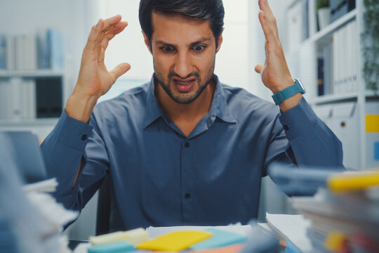 Closeup overworked young Latin businessman reading document in large stack of paperwork file at office desk. Tax audit deadline, accounting stress, corporate burnout, bookkeeping, bureaucracy concept.