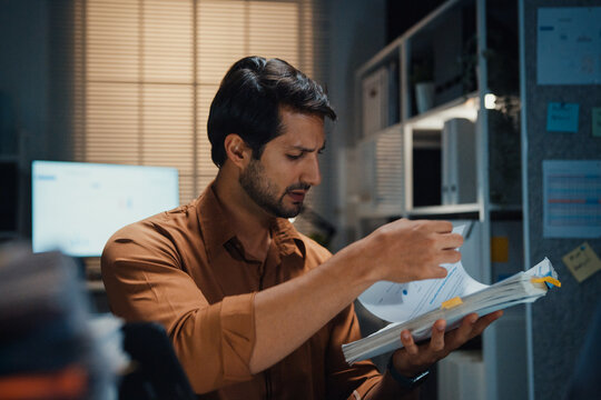 Overworked young Latin businessman working late on document in large stack of paperwork files at office night. Tax audit deadline, accounting stress, corporate burnout, urgent business concept.