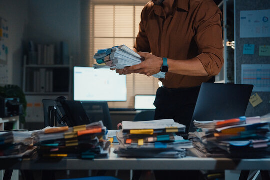 Stressed young Latin businessman walking to place tax document stack at desk in dark night office. Overworked overtime, heavy workload stress, tired employee, corporate business burnout concept.