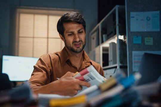 Stressed young Latin businessman having severe headache and migraine at desk in night office. Debt crisis, corporate burnout, late overtime workload, mental health anxiety, business failure concept.