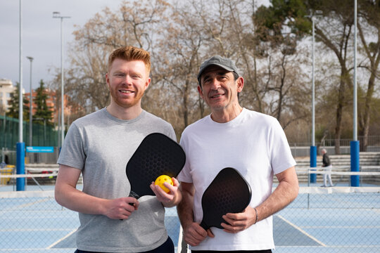 Two men smiling and holding pickleball paddles and a ball on an outdoor court