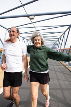 Happy senior man and woman jogging outdoors, smiling while exercising on a bridge
