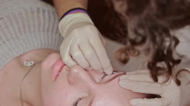 Beginner piercer attempts eyebrow piercing with visibly trembling hands, Unsteady gloved hands hold a needle near a young woman's eyebrow piercing