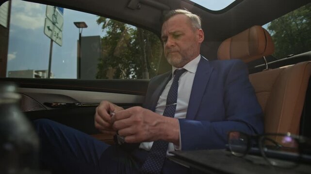 Middle aged Caucasian businessman sitting in luxury car, taking off glasses, rubbing forehead, and drinking water. Video is suitable for corporate wellness and executive stress campaigns
