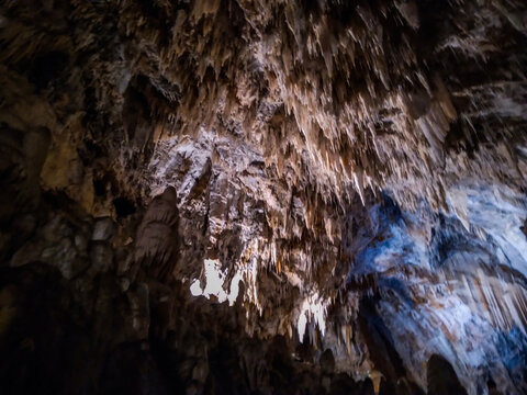 Massive jagged stalactites hanging from the dark rugged cavern ceiling showcasing ancient geological rock formations with blue lighting creating a mystical atmosphere inside this deep cave