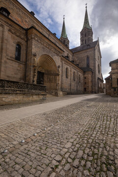 20 August 2026, View of the historic Bamberg Cathedral with its stone facade and spires along a cobbled street in Bamberg, Germany.