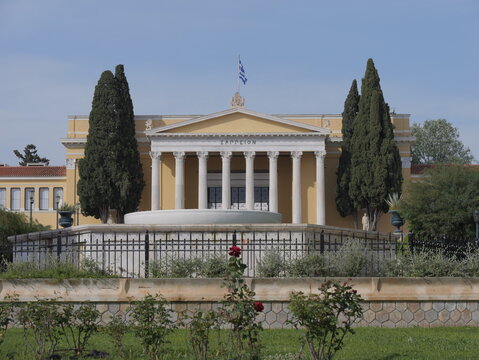 View of the Zappeion building with its neoclassical columns and pediment, featuring a fountain and lush rose bushes in the foreground under a clear blue sky in Athens, Greece.