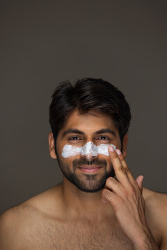 A YOUNG MAN HAPPILY APPLYING FACE CREAM WHILE LOOKING AT CAMERA