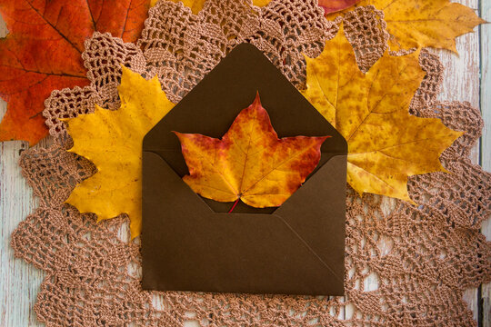 Cozy autumn flat lay with bright yellow and orange maple leaves in an open brown craft envelope on a hand-crocheted lace doily and wooden surface. Ideal for Thanksgiving cards.