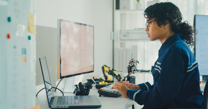 Young African American female robotics engineer coding AI software for robotic arm in laboratory. Embedded system, firmware testing, machine learning, industrial automation, future technology concept.