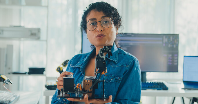 Young African American female robotics engineer discussing mechanical part on video call and looking at camera in laboratory. Remote work, virtual training, online meeting, future technology concept.