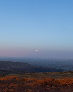 View of the moon rising over the rolling hills and rocky terrain of Bannau Brycheiniog National Park at dusk in Brecon, Wales, United Kingdom.