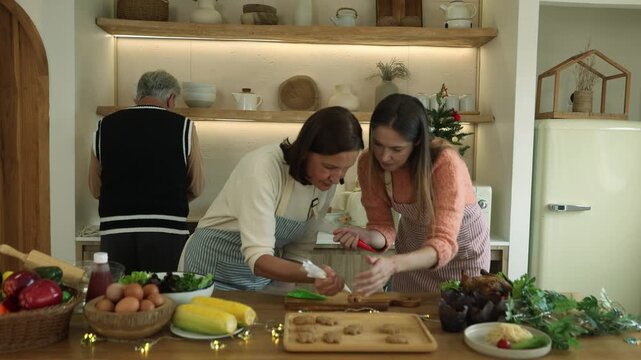 Mother and grown up daughter are preparing decorate baked cookies on the kitchen counter getting ready for the family Christmas party while the father stands behind them washing vegetables.