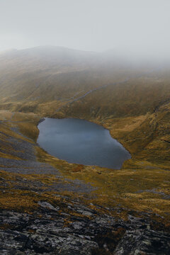 View of the mountain tarn below Sharp Edge, surrounded by grassy slopes and rocky terrain under a misty sky in Keswick, England, United Kingdom.