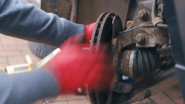 Gloved hands attempting removing seized old brake disc from wheel hub. Automotive repair struggling with rusted brake rotor during car maintenance process. Closeup manual disassembly with hydraulic