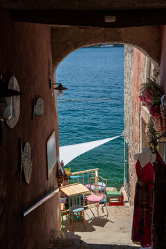 View of a charming stone archway leading to a seaside cafe with colorful chairs and a mannequin displaying a dress overlooking the Adriatic Sea in Rovinj, Istria County, Croatia.