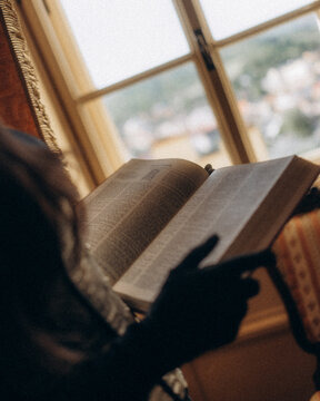 View of a person reading a book by a window with a blurred view of a town in the background CZ.