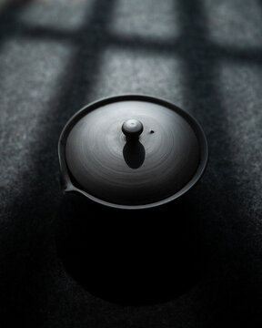 View of a black ceramic teapot with a lid resting on a dark textured surface with dramatic grid shadows CZ.