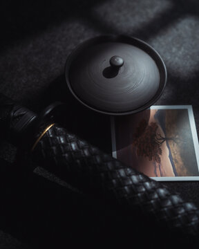 View of a black katana handle, a dark ceramic bowl, and a small photograph of a sunset landscape on a dark surface CZ.