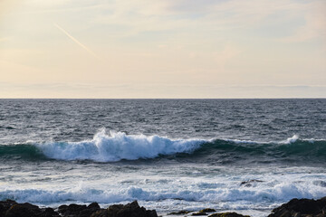 Fototapeta premium Powerful ocean wave breaking towards a rugged rocky shoreline beneath a soft atmospheric sky, with natural sea textures and generous copy space. A versatile coastal seascape for travel, nature, weathe