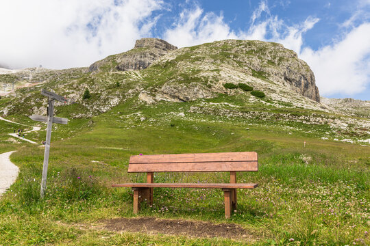 Wooden bench facing a mountain with a grassy path under a partly cloudy sky. Dolomites, Italy