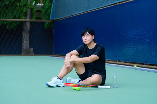 Young Asian Male Tennis Player Resting on Court with Racket and Water Bottle After Practice