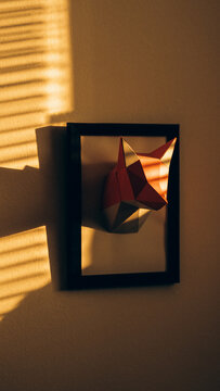 View of a low-poly paper fox head sculpture mounted in a black frame on a wall, illuminated by warm sunlight casting striped shadows in Svitavy, CZ.