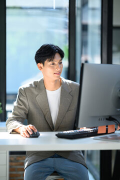 Young Asian Businessman Working on Desktop Computer at Modern Office Desk with Natural Light