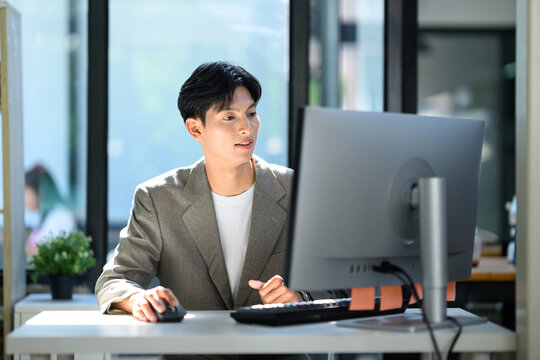 Young Asian Businessman Working on Desktop Computer at Modern Office Desk with Natural Light