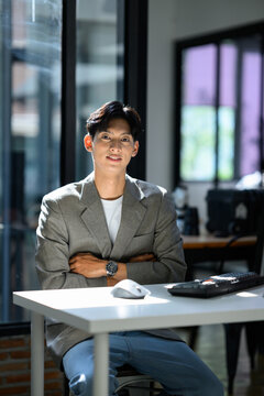 Young Asian Businessman Sitting at Office Desk with Arms Crossed, Confident Smile, Gray Blazer