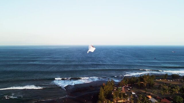 Vibrant long tail kite soars in the wind over foaming sea waves with colorful textile patterns stretching across a clear open horizon and vast ocean below aerial drone footage