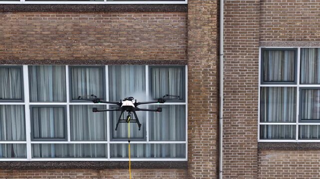 Autonomous drone with a tethered hose and sprayer conducting high pressure washing on the windows of a large brick building, representing the future of industrial cleaning technology