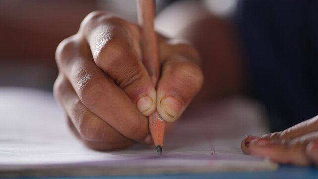 Close-up of a student's hand gripping a pencil while writing on white paper in rural India. Shallow depth of field isolates the writing hand against a soft-focused background.