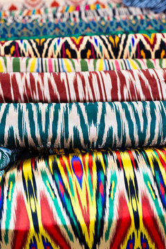 Colorful Uzbek ikat fabrics stacked at a bazaar in Bukhara. Traditional silk textiles with vibrant patterns displayed for sale. Ideal for travel, culture, and textile design content.