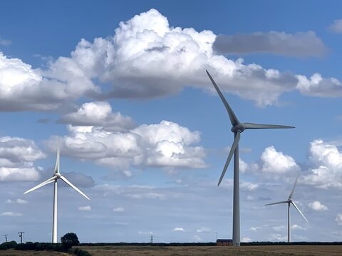 View of Burton Wold wind farm with three large white wind turbines standing in a field under a bright blue sky with fluffy white clouds in Burton Latimer, England, United Kingdom.